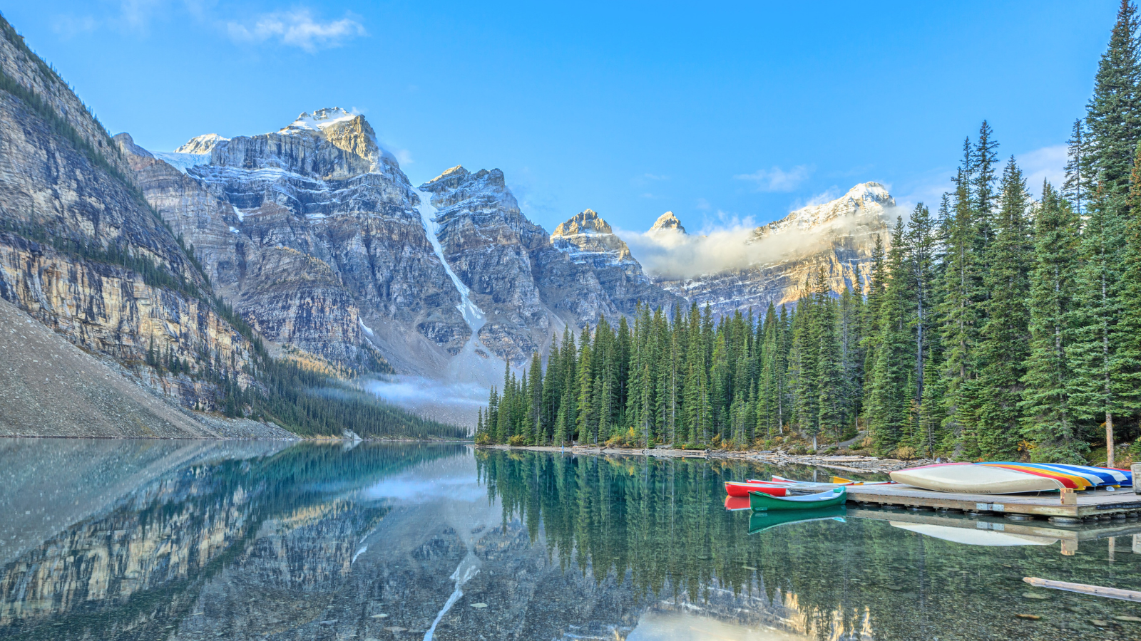 Mountains reflect in a mirror-like lake, with kayaks on a jetty and a forest to the side